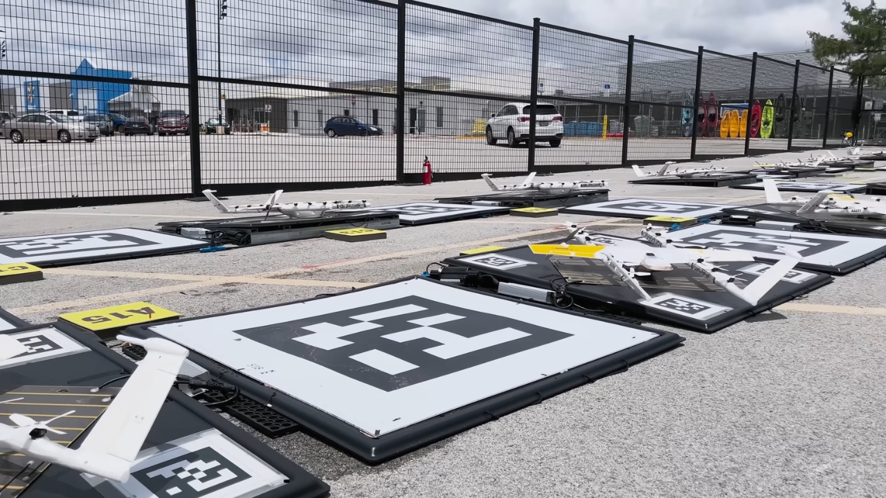 Several delivery drones sit on launch pads inside a fenced outdoor facility.