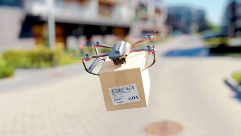 A delivery drone carries a cardboard package above a street in a residential neighborhood.
