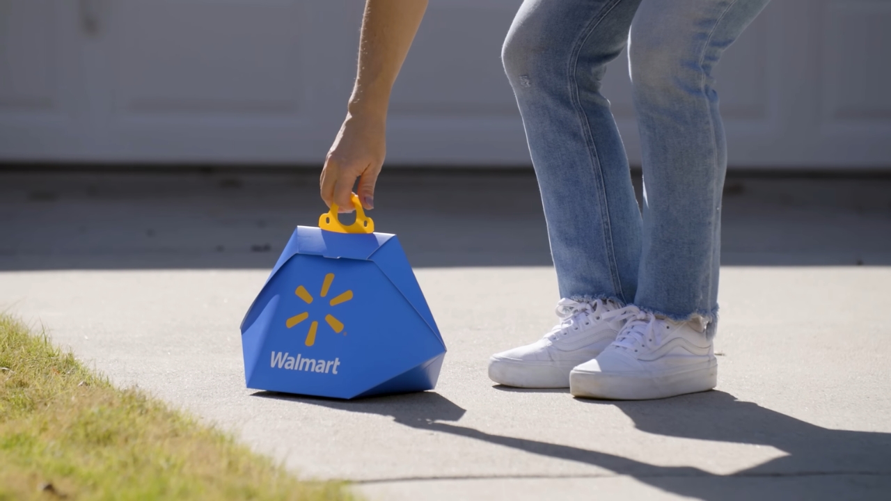 A person picks up a small blue Walmart delivery package from a driveway.