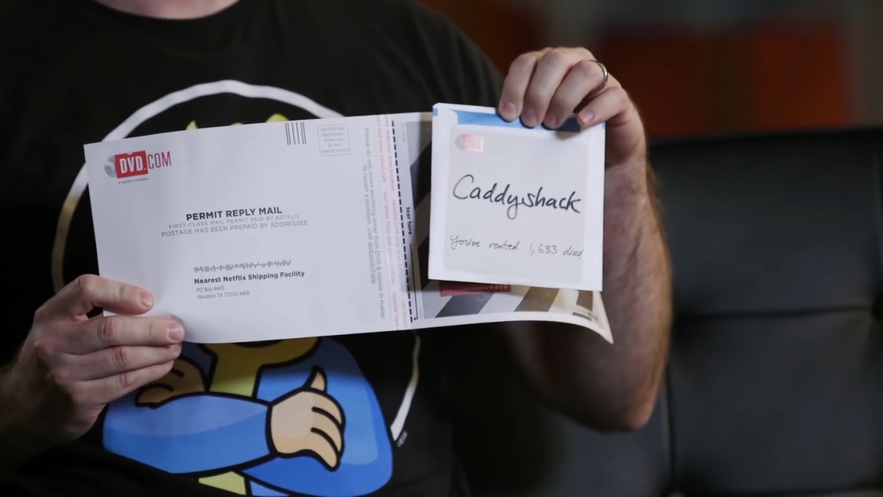 A person holds a Netflix DVD return envelope and a Caddyshack DVD sleeve.