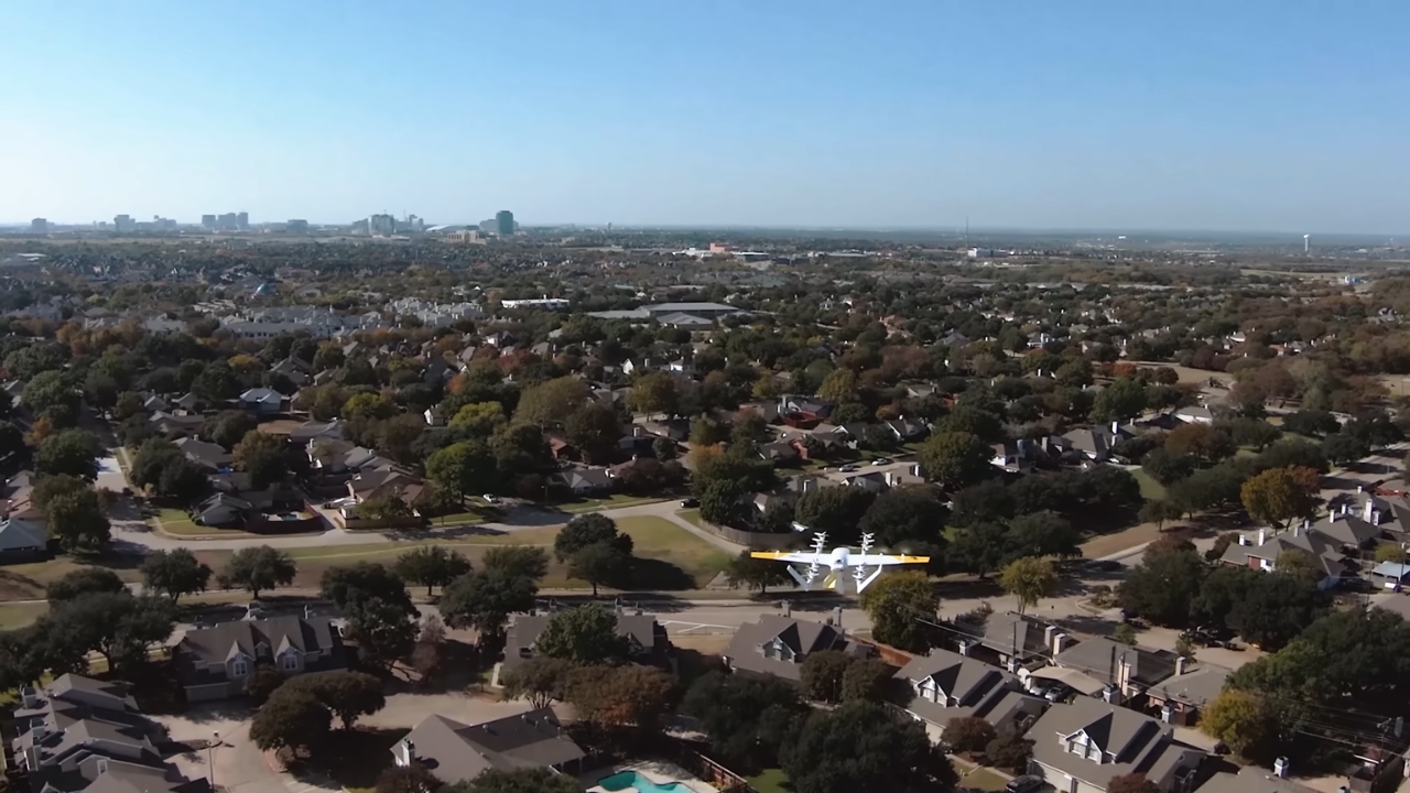 A delivery drone flies above a suburban neighborhood with houses and tree-lined streets.
